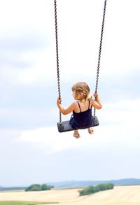 Rear view of girl on swing at playground against cloudy sky