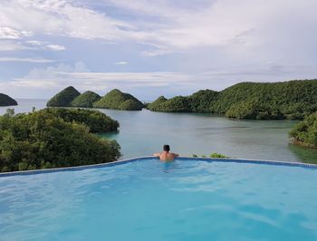 Rear view of man swimming in infinity pool by sea against sky