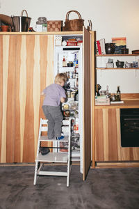 Rear view of boy standing on ladder and searching in refrigerator at home