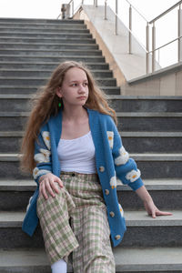 A young girl, blonde with long hair, in a blue cardigan is sitting on the steps of a gray staircase