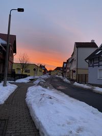 Snow covered road by buildings against sky during sunset