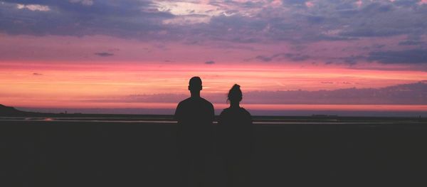 Rear view of silhouette man and woman standing on landscape against orange sky