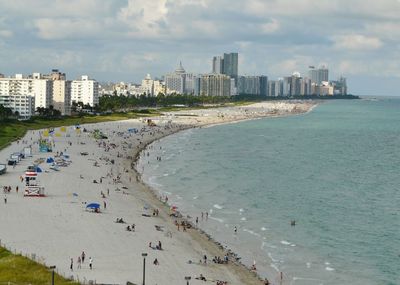 Aerial view of beach and buildings against cloudy sky during sunny day in city