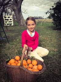 Portrait of girl holding basket with oranges while crouching on grassy field