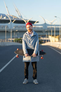 Young bearded hipster man in red hat hold longboard in city street. trendy urban lifestyle concept