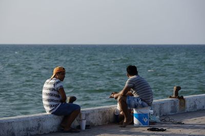 Rear view of boys sitting on shore against clear sky