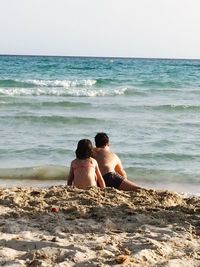 Rear view of shirtless man sitting on beach against clear sky