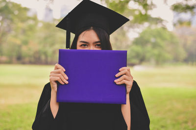 Portrait of young woman in graduation gown holding file while standing at park