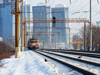 An old electric train moves on rails in the winter season against the backdrop of a cityscape.