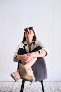 Full body of positive female in trendy outfit and high heels sitting on rug near white wall in light studio