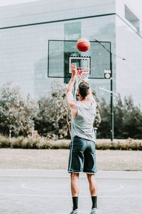 Full body back view of unrecognizable determined young male basketball player in sportswear leaping and shooting ball near hoop while training alone on street playground