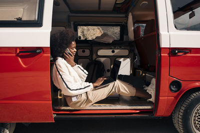 Side view of young woman talking on smart phone while using laptop sitting in van during vacation