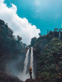 Man standing on rocks by waterfall against sky