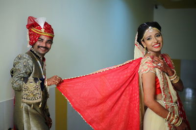 Smiling groom holding sari of bride while standing against wall at home