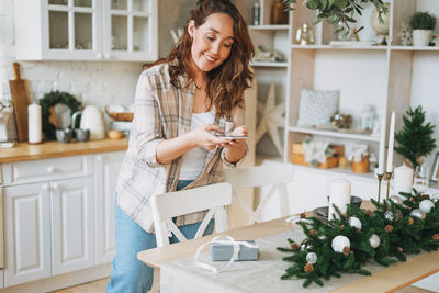 Attractive curly hair woman in plaid shirt takes photo gift box on festive table 