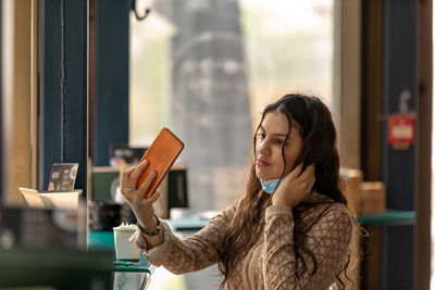 Cute teen wearing medical mask sitting in cafe touching and combing hair using smartphone as mirror