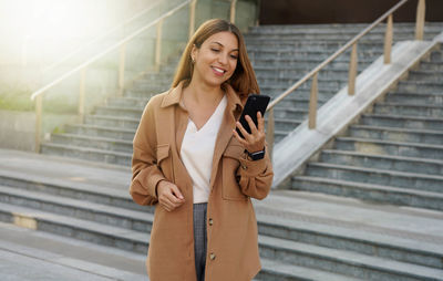 Successful businesswoman holding mobile phone while walking outdoors near financial office