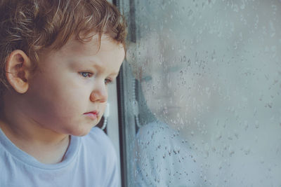 Sad child looking out the window with wet glass autumn bad weather.