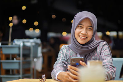 Beautiful asian muslim woman wearing modern hijab sitting smiling and holding smartphone in a cafe