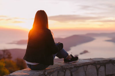 Woman sitting on retaining wall against sky during sunset