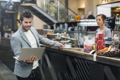 Mid adult man holding laptop while buying coffee at cafe