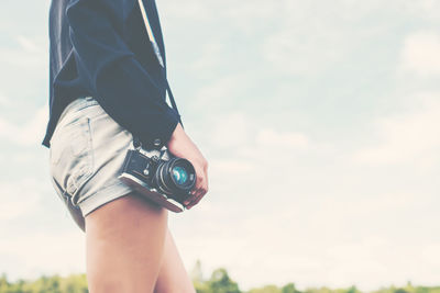 Midsection of woman with camera standing against sky