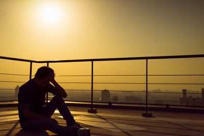 Side view of woman sitting on railing against sky during sunset