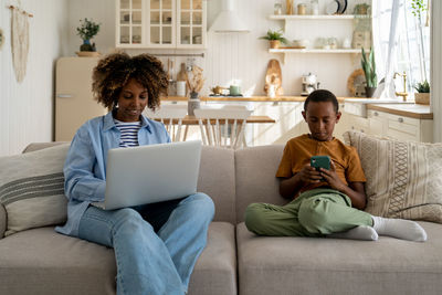 African mom freelancer remote work from home office on laptop sit on couch child playing on phone