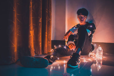 Portrait of young man with illuminated string lights while sitting against wall
