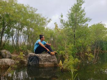 Man sitting on rock in lake against trees and sky