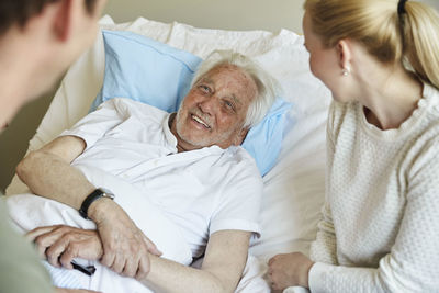 Happy senior man talking to couple while reclining on bed in hospital ward