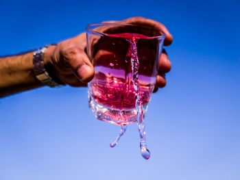 Cropped hand of man pouring drink from glass against clear blue sky