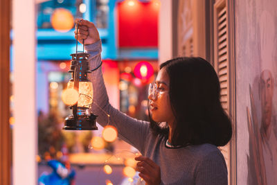 Side view of woman looking at illuminated lantern while sitting in corridor