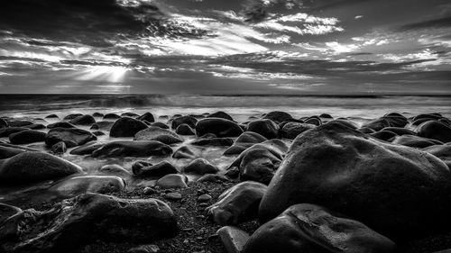 Black and white photo of stony beach with oceanside background beneath stormy sky