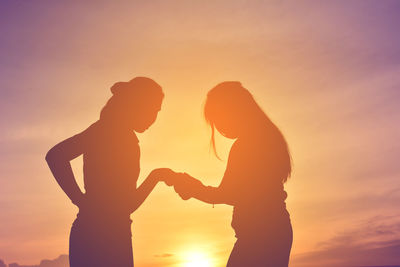 Silhouette female friends holding hands while standing against orange sky