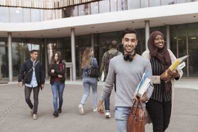 Portrait of smiling student holding books while walking in campus with friends in background