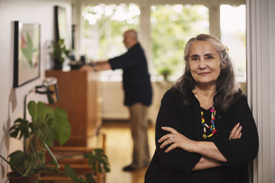 Portrait of confident senior woman standing with man in background at home