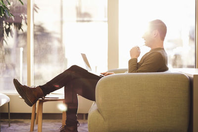 Full length of businessman using laptop while sitting on sofa during sunny day