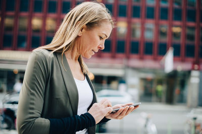 Blond mature businesswoman using smart phone against building in city