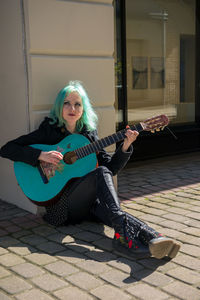 Hipster young woman playing turquoise guitar while sitting on street in city