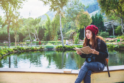 Young woman reading book while sitting by pond