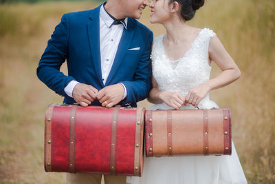 Midsection of bridal couple holding suitcases on field