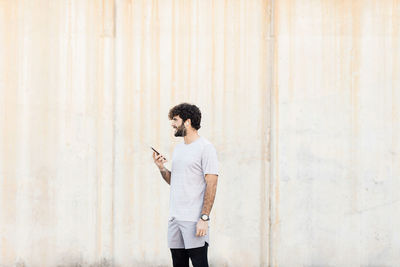 Full length of young man using phone while standing against wall