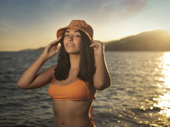 Portrait of young woman in bikini standing in sea against sky during sunset