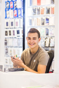 Portrait of smiling young male trainee repairing mobile phone while sitting at illuminated desk in store