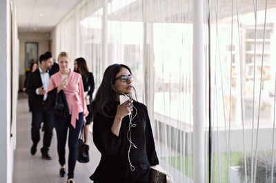 Businesswoman talking on smart phone in corridor while leaving office with colleagues in background