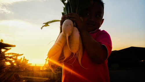 Low angle view of boy holding radish while standing against sky during sunset