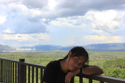 Portrait of young woman standing on railing against sky