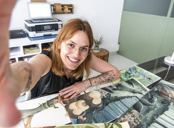 Cheerful female designer sitting at table with art supplies and taking self shot on smartphone in workspace