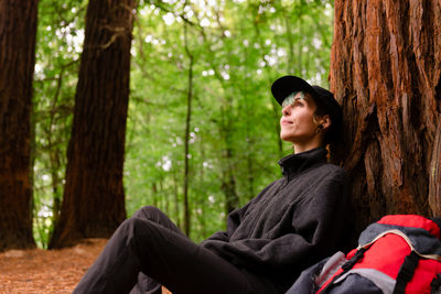 Delighted explorer with backpack sitting near huge tree in forest and resting during trekking in monte cabezon natural monument of sequoias while looking up